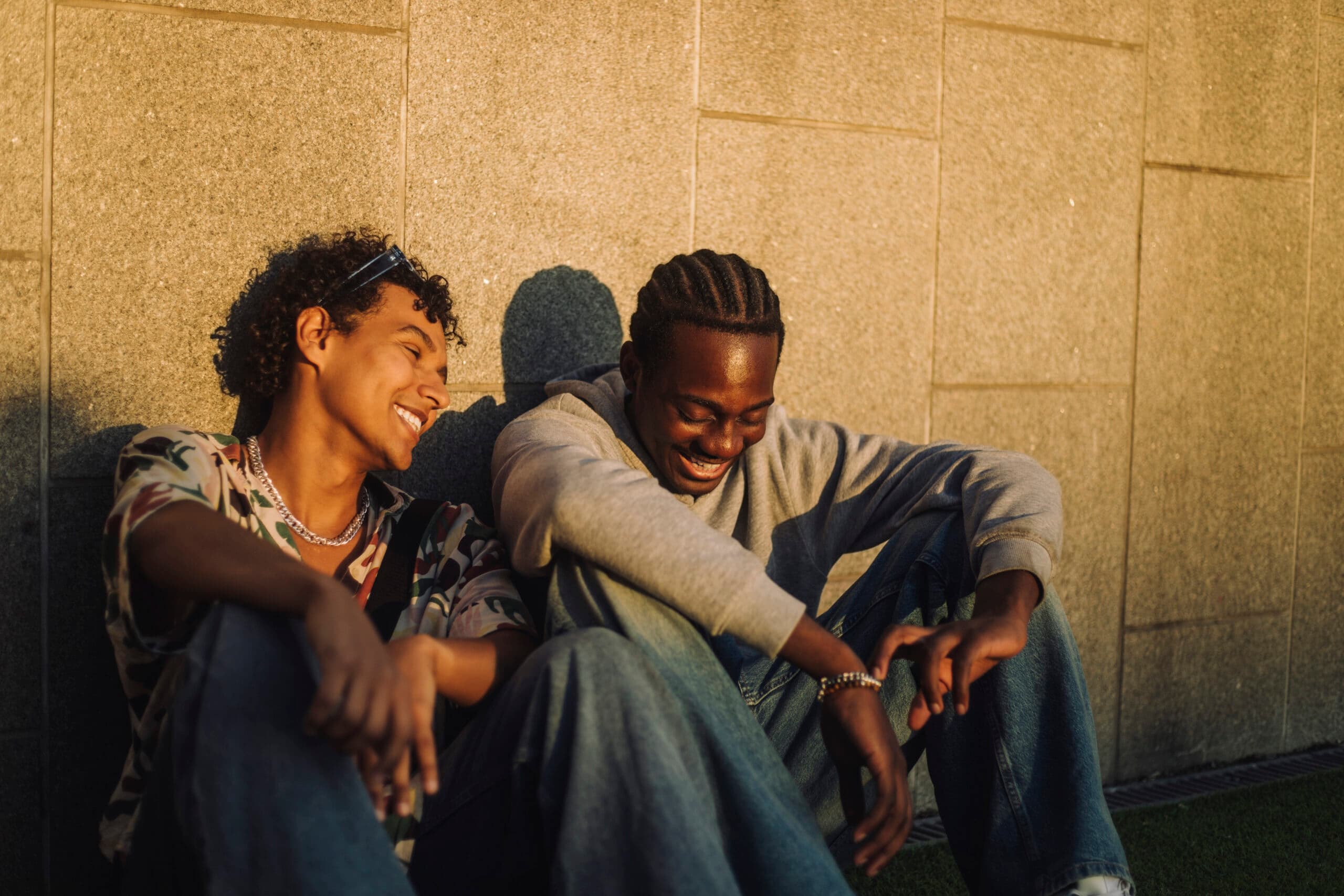 Happy male friends sitting near wall at sunset smiling together.