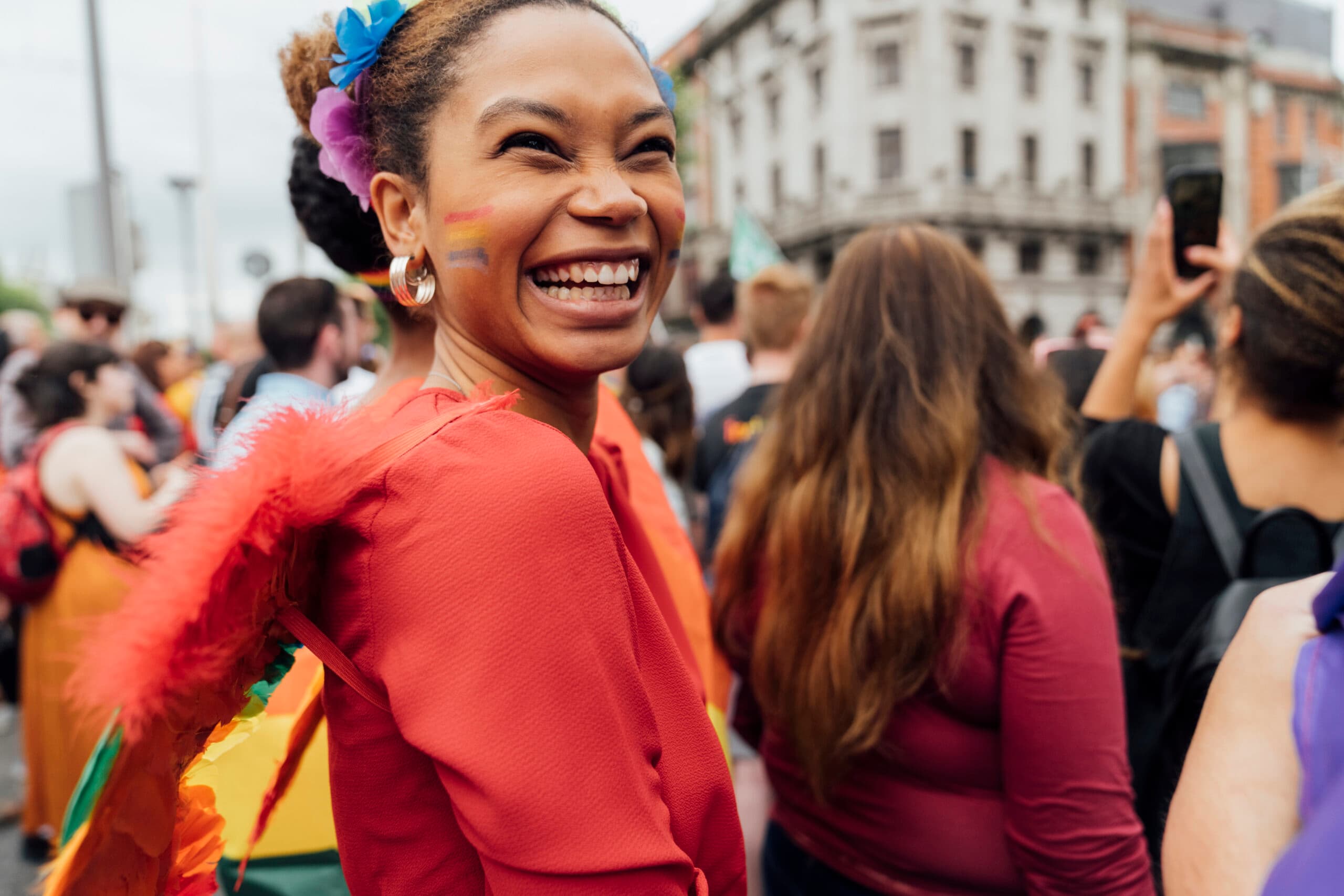 Positive and Candid Smile of a Black Afro Woman standing Among the People . She is Wearing a Summer Red top and Rainbow Colours for a great Celebration of the Gay Pride
